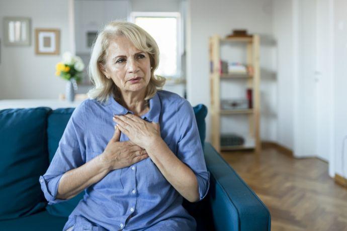 Eine ältere Frau sitzt mit Schmerzen in der Brust auf dem Sofa.