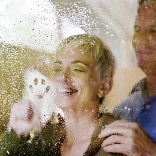 Ein lächelndes Paar schaut durch ein feuchtes, beschlagenes Fenster, während die Frau innen einen Smiley-Gesicht aufmalt.