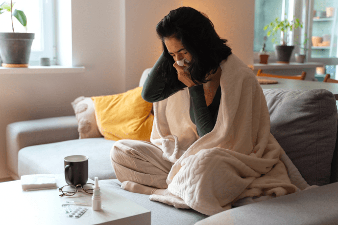 Eine Frau sitzt in eine Decke gehüllt auf dem Sofa und putzt sich die Nase, weil sie krank ist.
